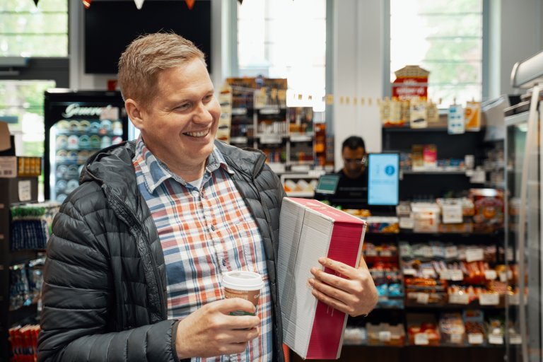 En mand står ved en pakkeshop med en pakke i den ene hånd og en kop kaffe i den anden