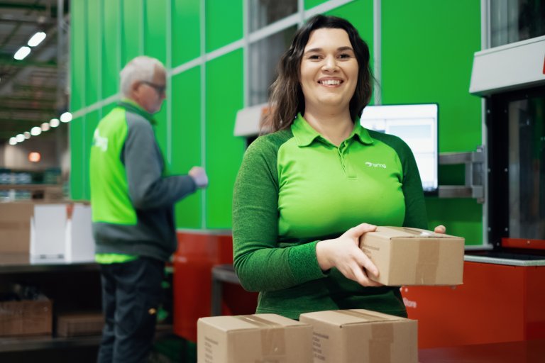 En smilende kvinde holder en pakke i hånden, mens hun arbejder i Autostore